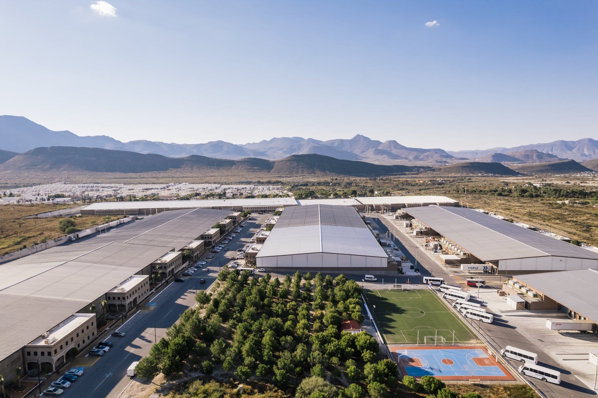 Aerial view of a Tetakawi manufacturing campus in Saltillo, Mexico, showing warehouse facilities, employee soccer field, basketball court, and shuttle buses | Tetakawi