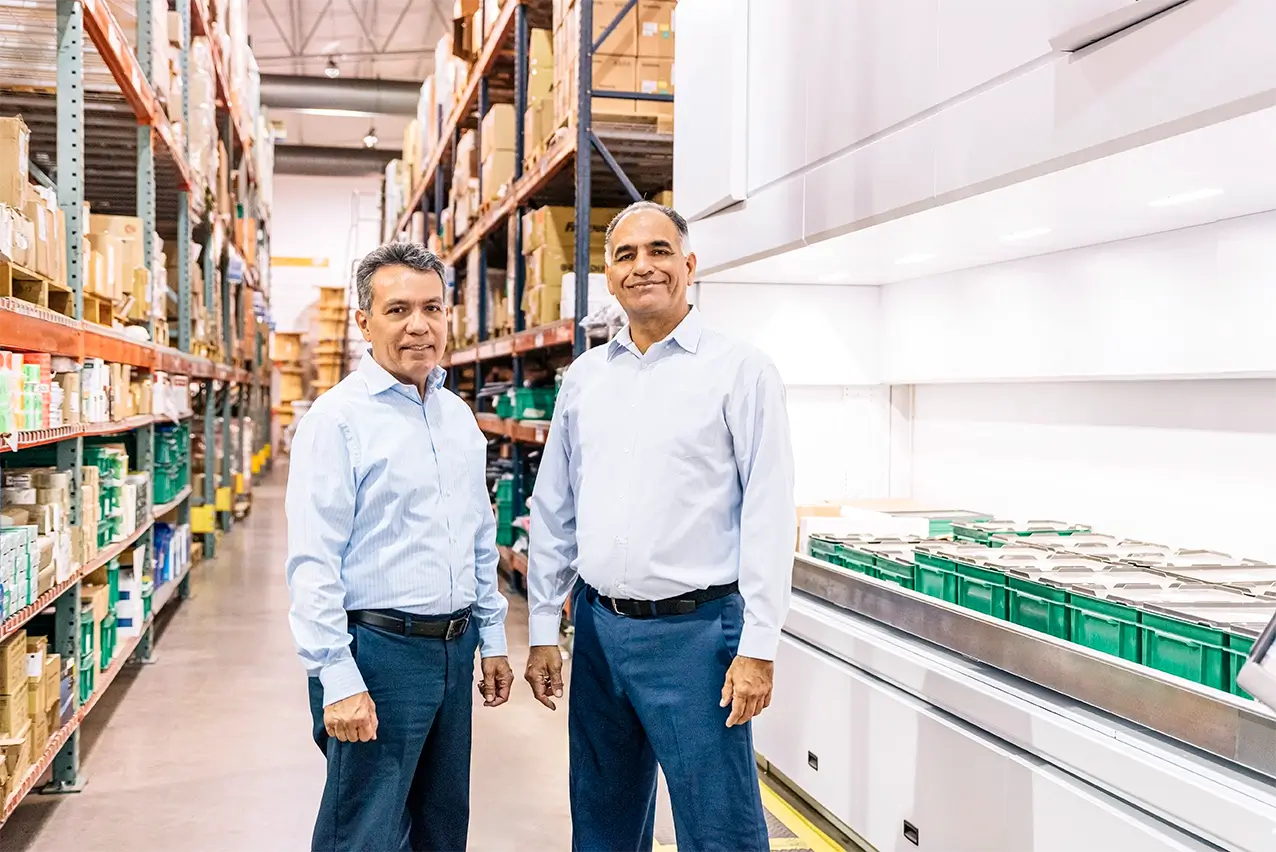 Two managers in light blue shirts standing beside inventory racking and automated bins, manufacturing in Mexico | Tetakawi