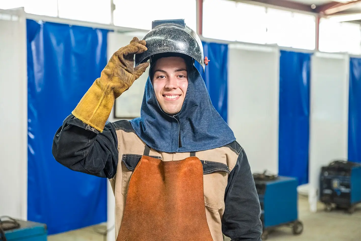 Smiling welder in a leather apron lifting his helmet beside welding booths, manufacturing workforce in Mexico | Tetakawi