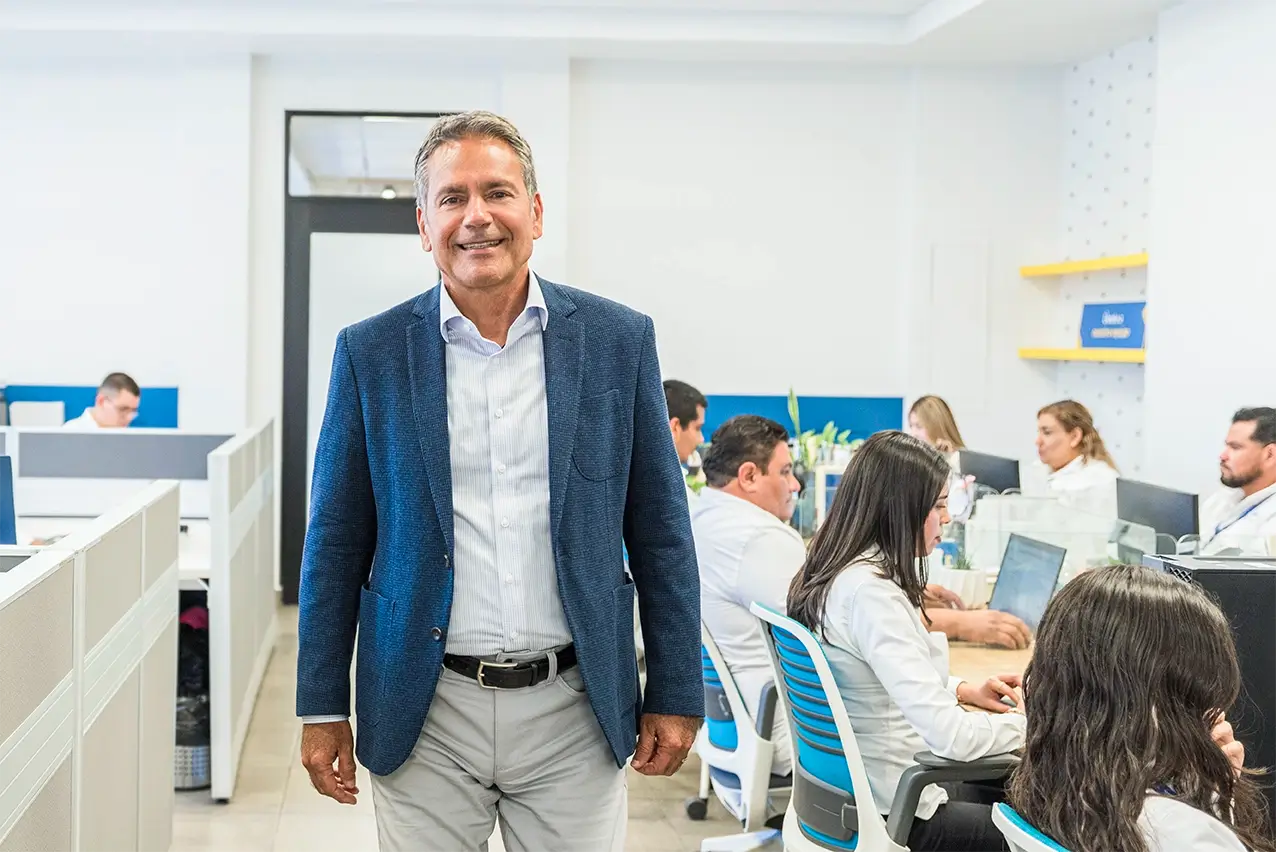 Smiling executive in a blue blazer standing in an open office with staff working at desks, manufacturing in Mexico | Tetakawi