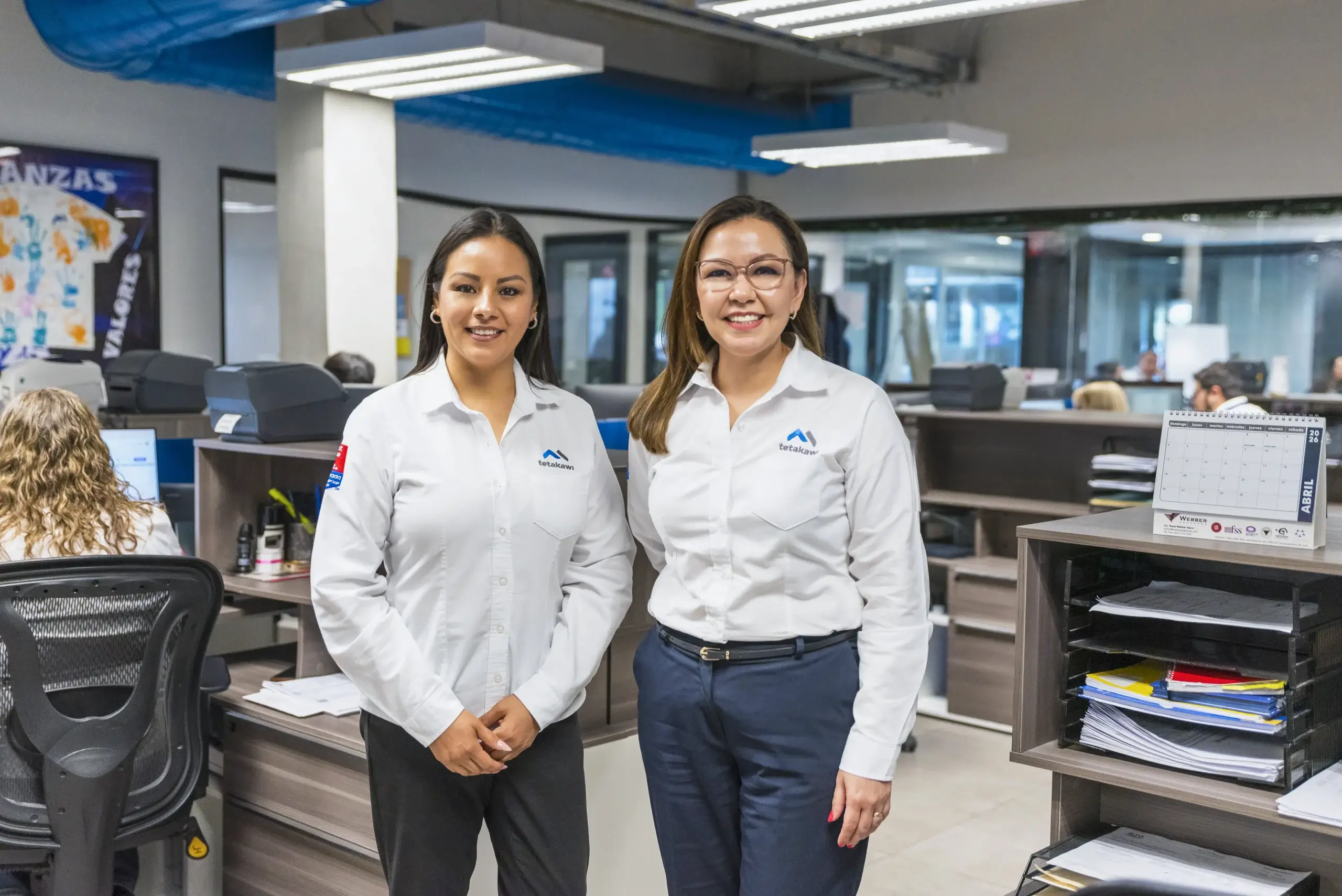 Two Tetakawi team members in branded white shirts standing in an open office, supporting manufacturing in Mexico | Tetakawi
