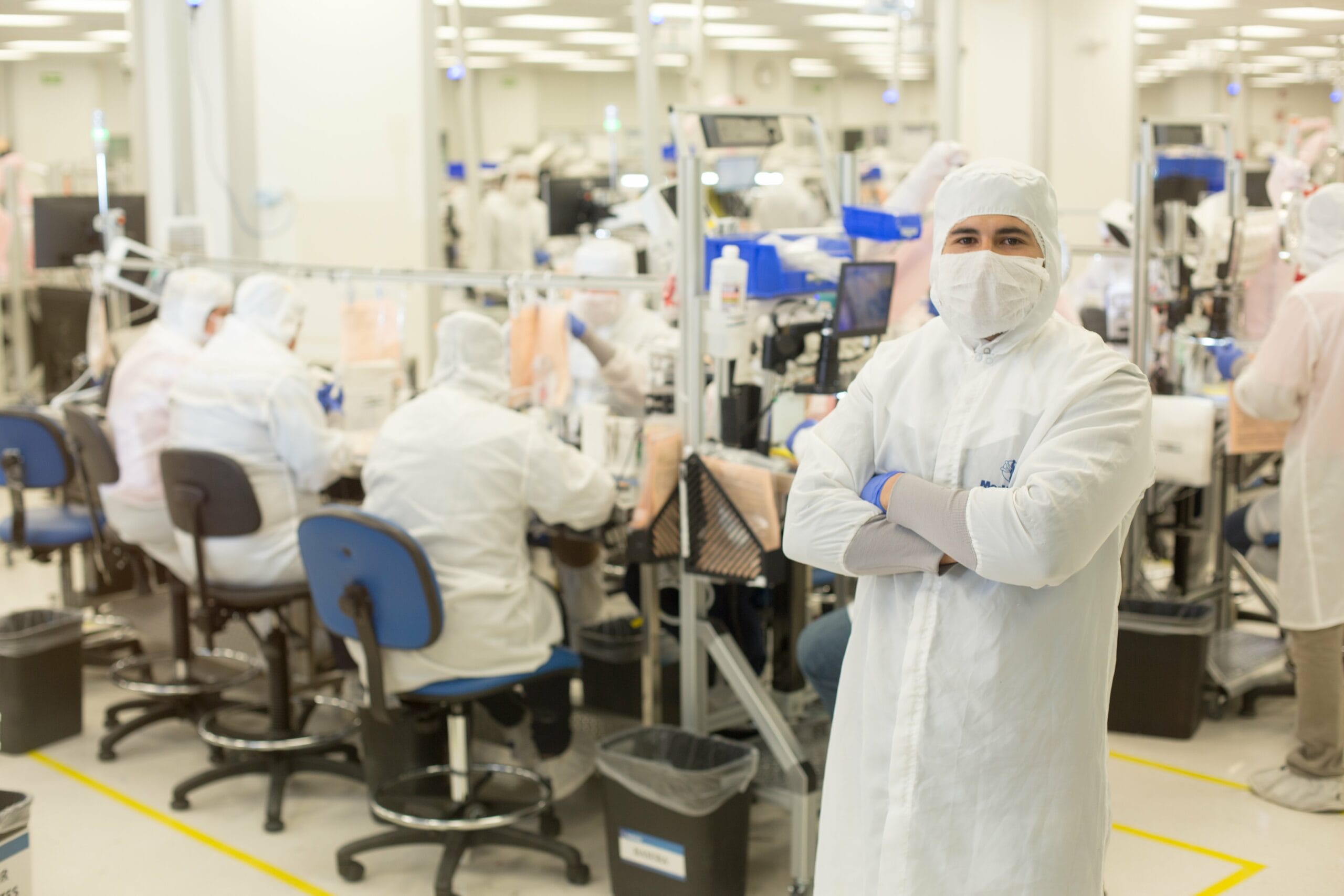 A technician in cleanroom PPE stands over a busy medical device assembly line in Mexico | Tetakawi