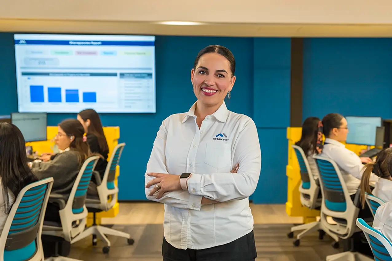 Smiling Tetakawi team member in an operations room with staff and a dashboard screen, manufacturing in Mexico | Tetakawi