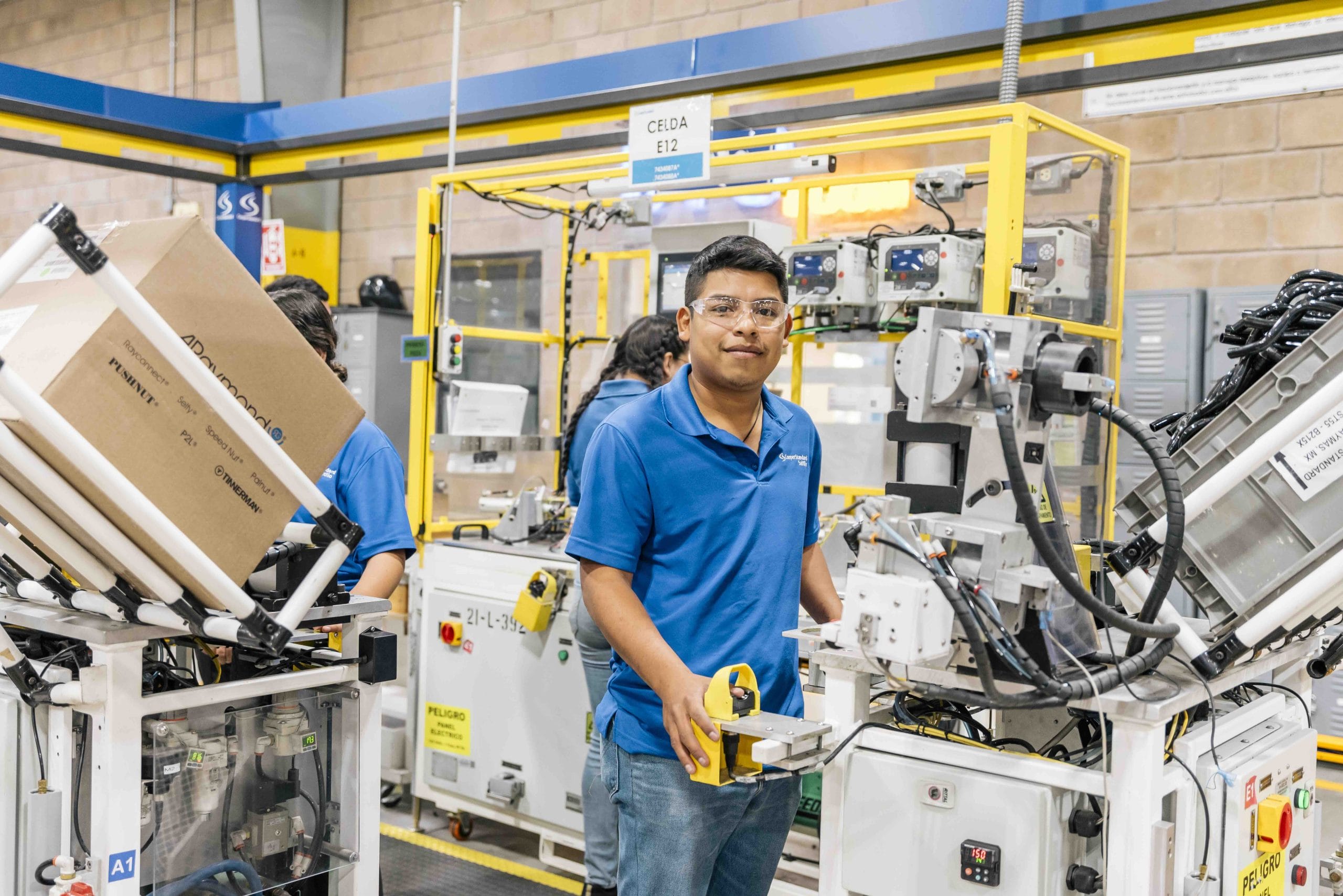 Production operator in safety glasses holding a fixture beside automated assembly cells, manufacturing in Mexico | Tetakawi