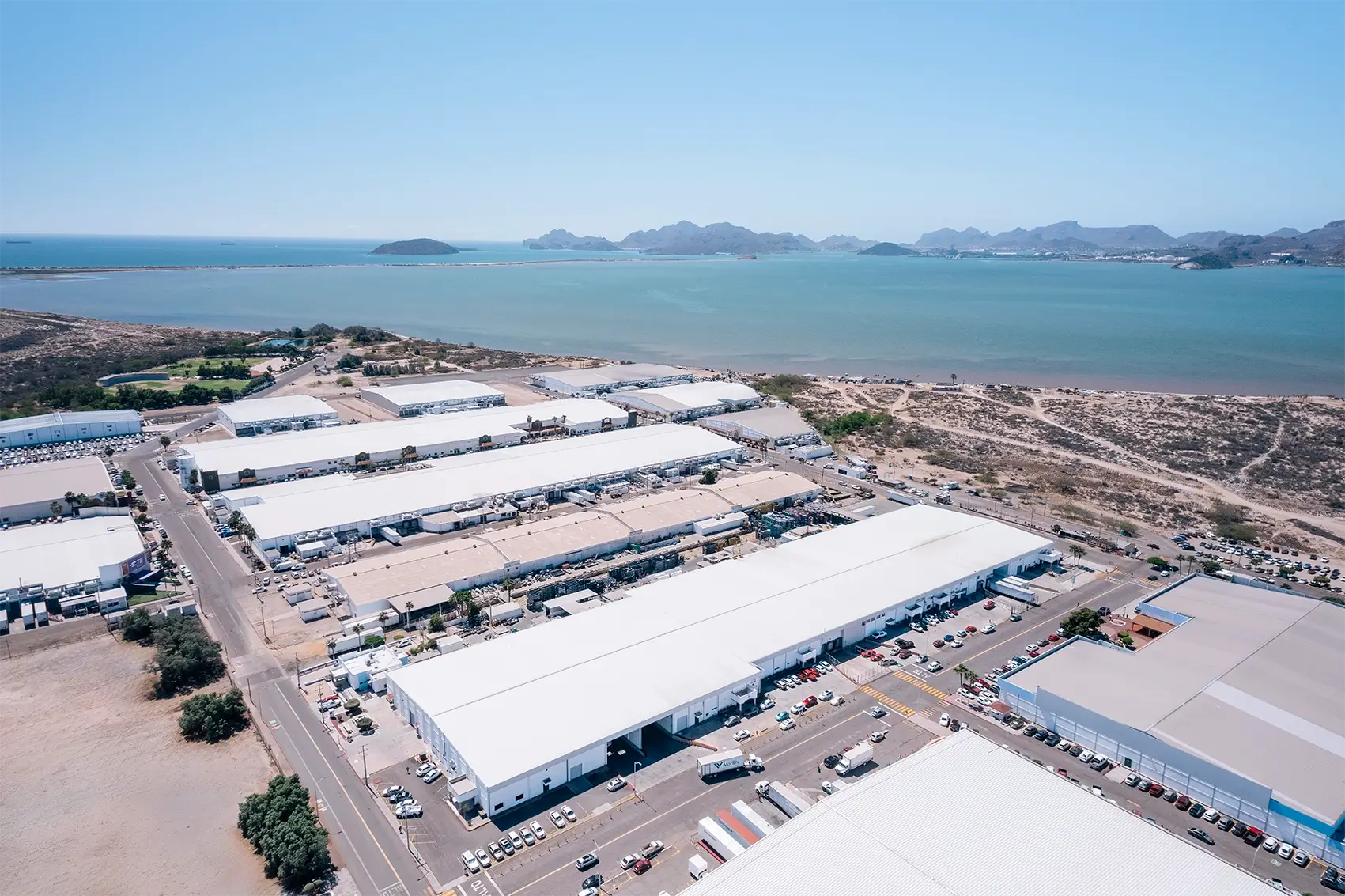 Aerial view of a Tetakawi Manufacturing Campus with white-roofed industrial buildings beside a bay in Mexico | Tetakawi