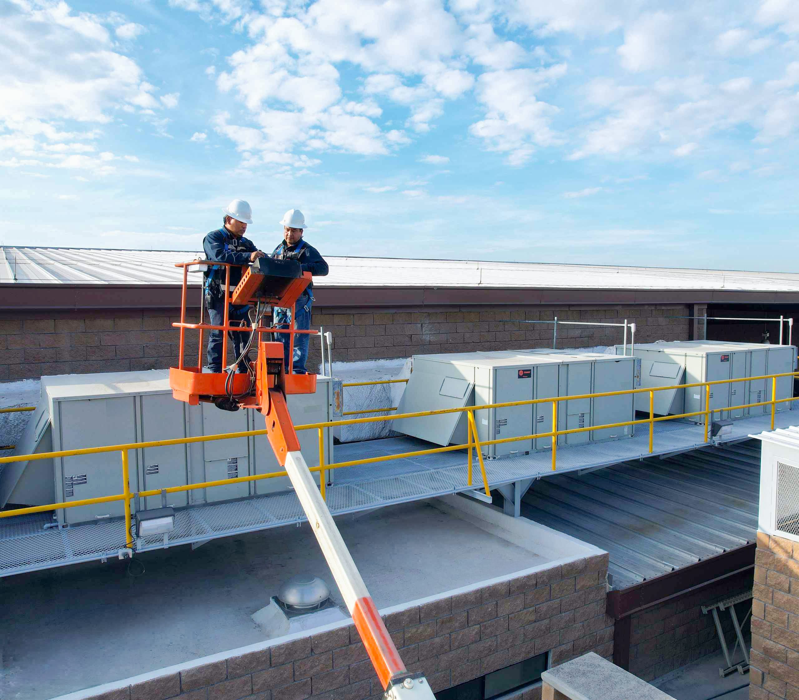 Two Tetakawi facilities technicians in hard hats on a lift servicing rooftop air-conditioning equipment at a Manufacturing Campus in Mexico