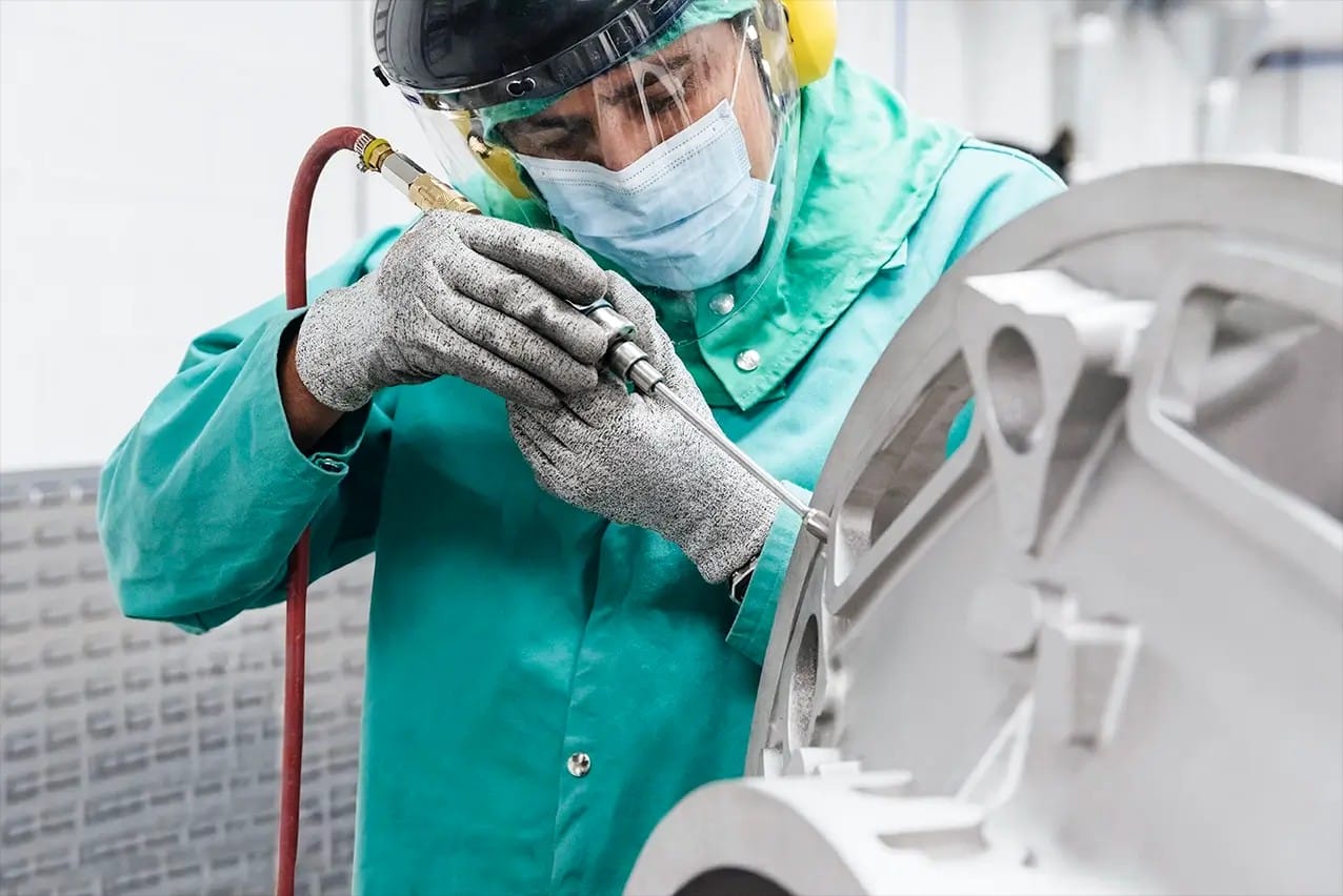 Technician in a face shield and protective coat finishing a large metal casting at a manufacturing plant in Mexico | Tetakawi